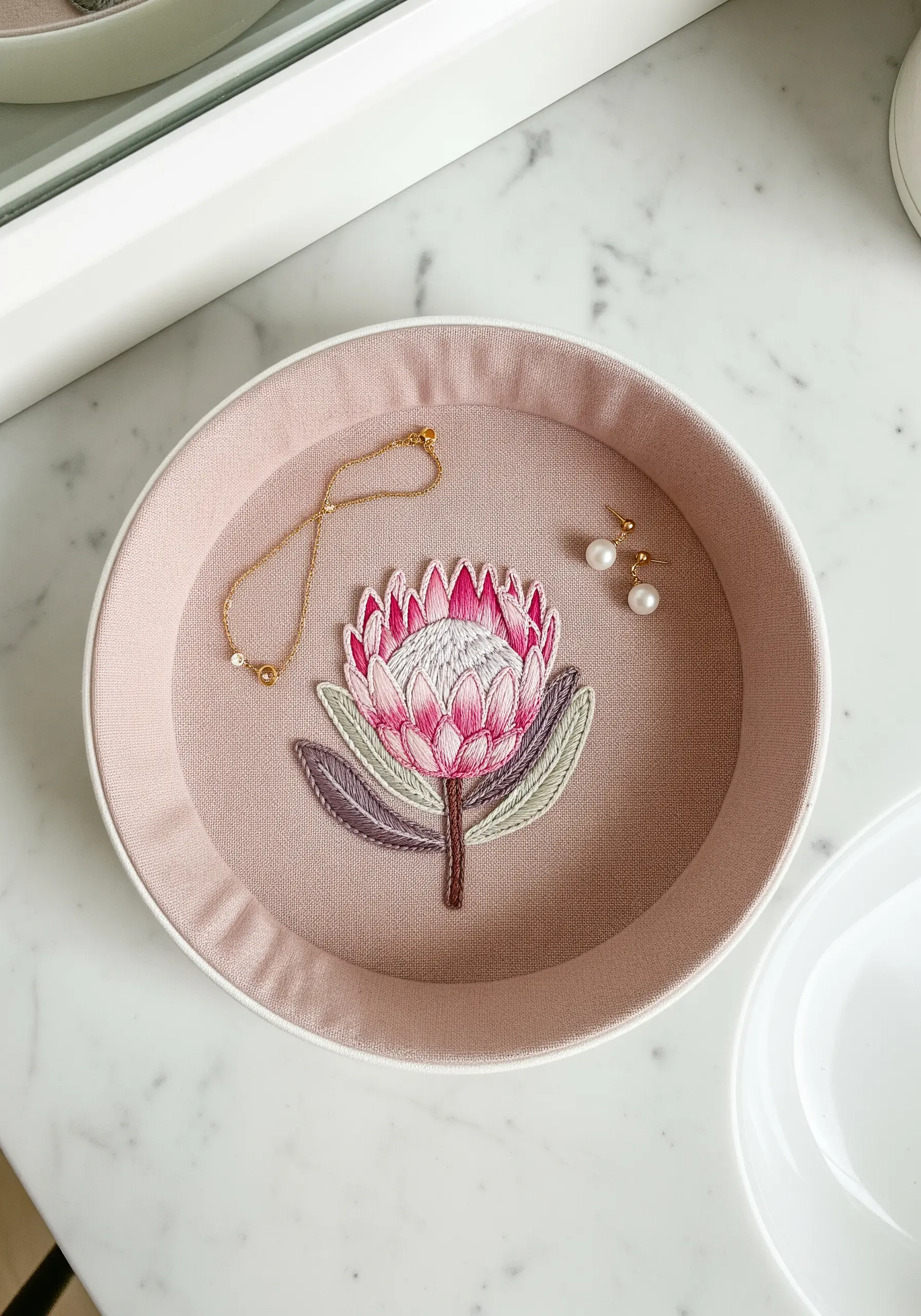 An embroidered protea flower in a pink fabric-wrapped hoop used as a jewelry tray.