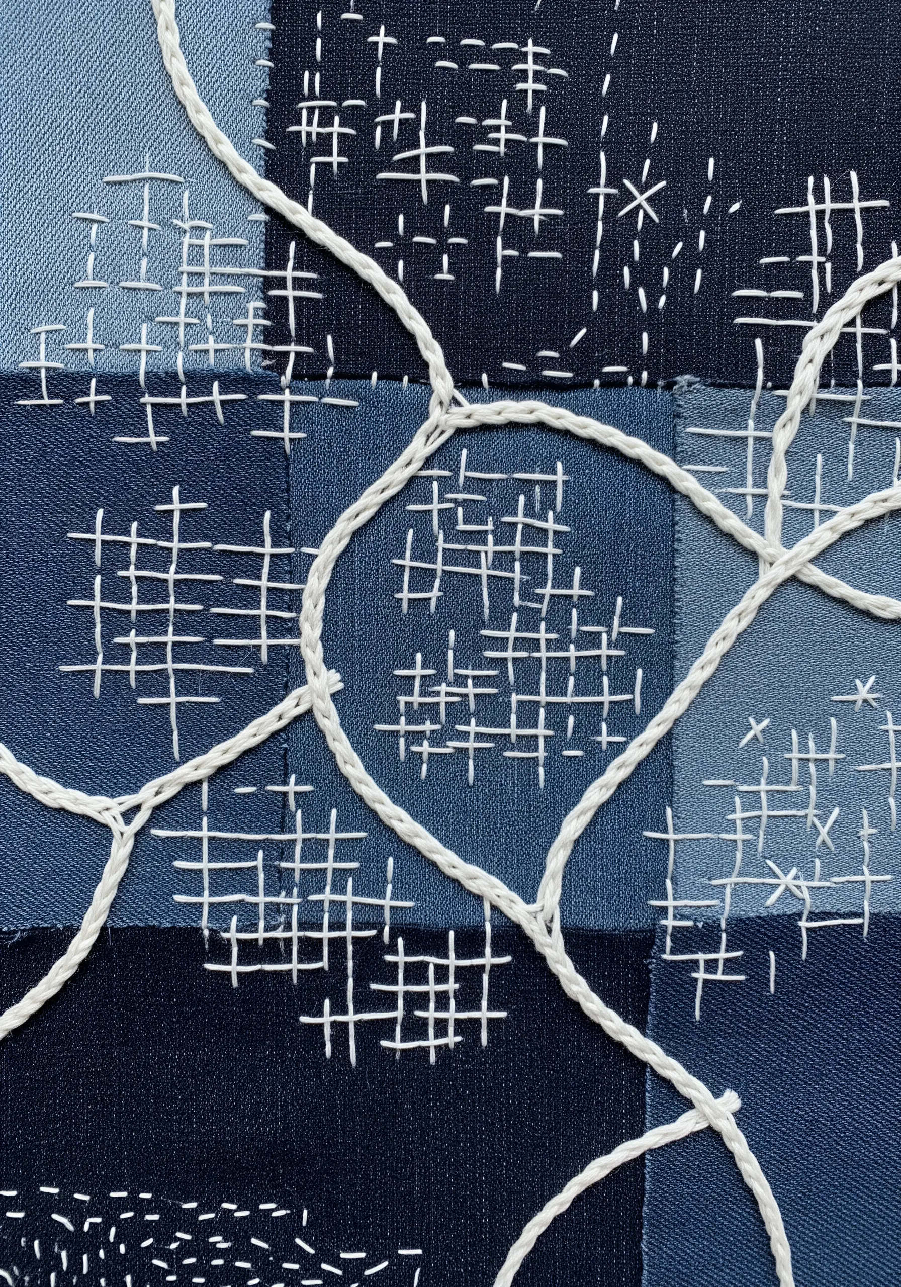 A patchwork of blue denim squares unified by a network of white running stitches and couched cord.