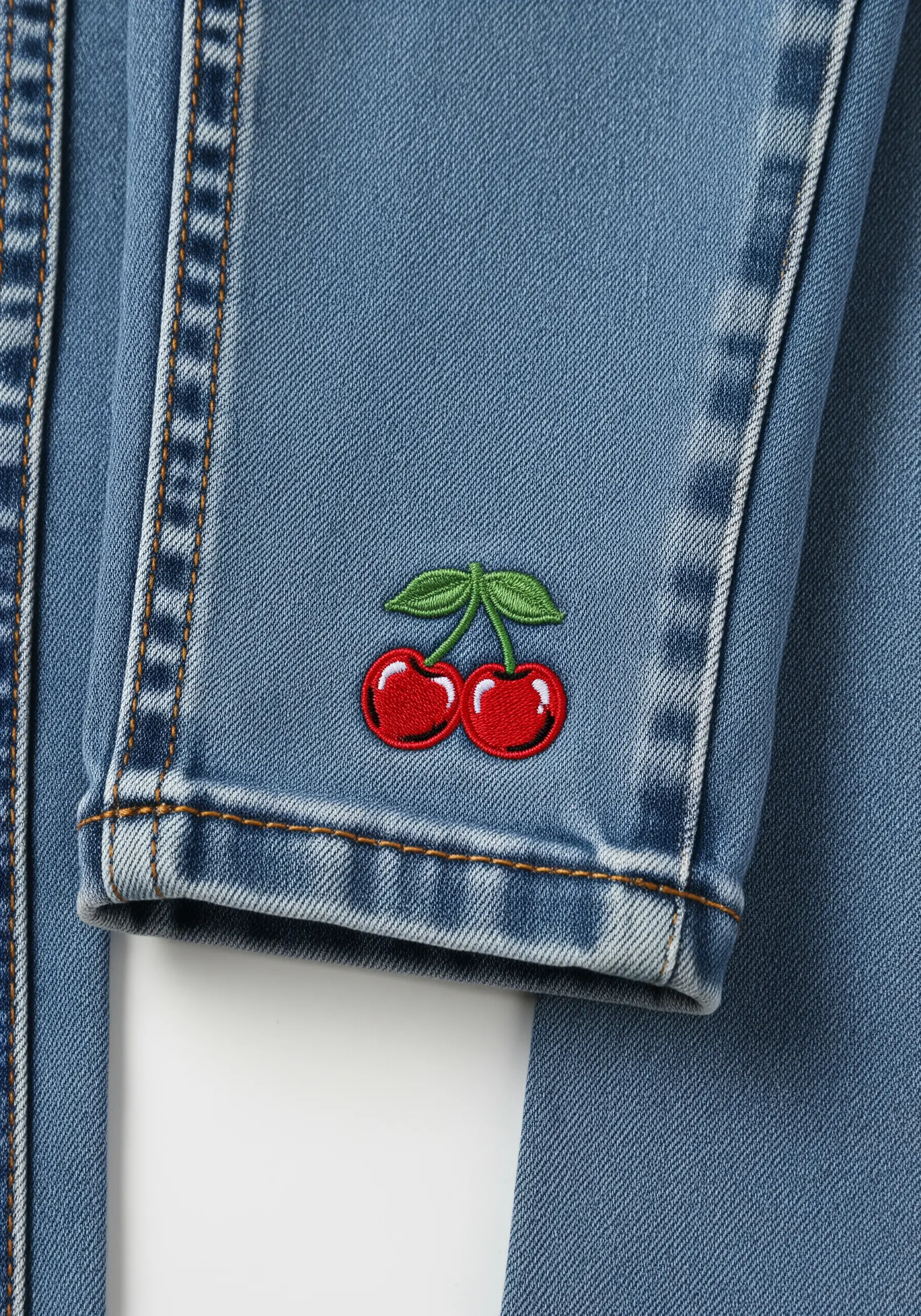 A pair of bright red embroidered cherries with a white highlight, stitched on a denim cuff.