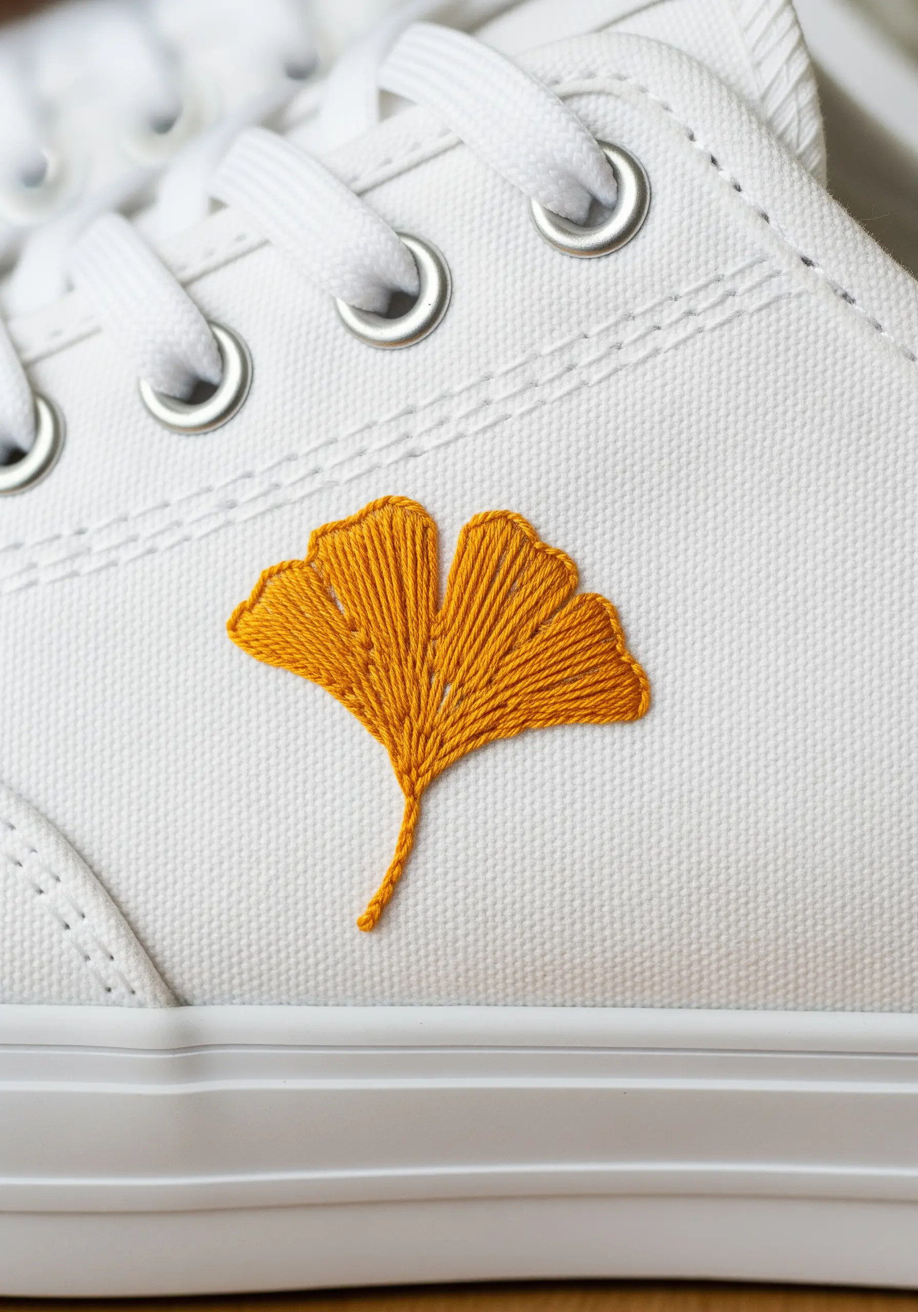 A golden-yellow ginkgo leaf embroidered on the side of a white canvas sneaker.