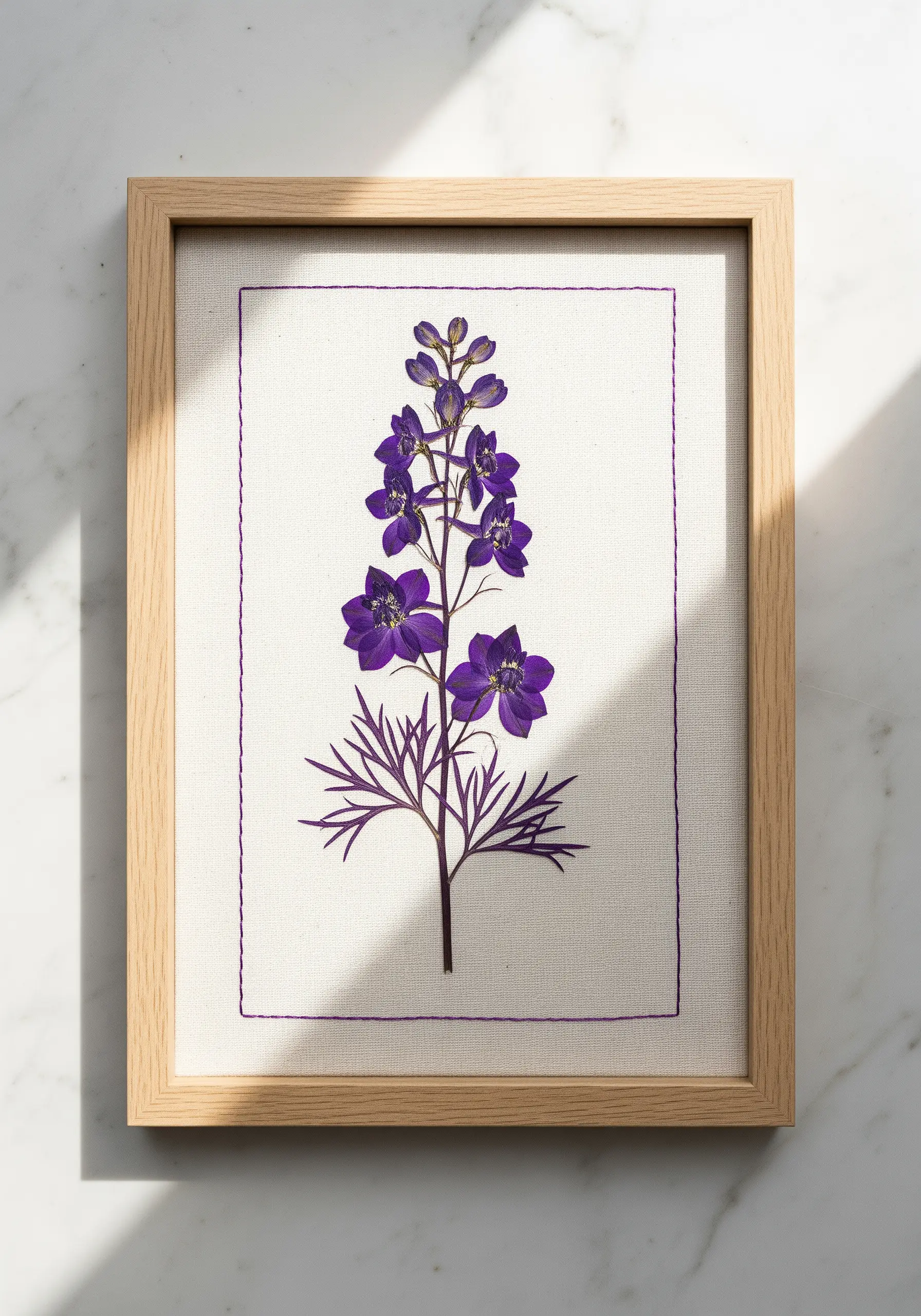 A single pressed delphinium on linen canvas, enclosed by a simple purple running stitch border.