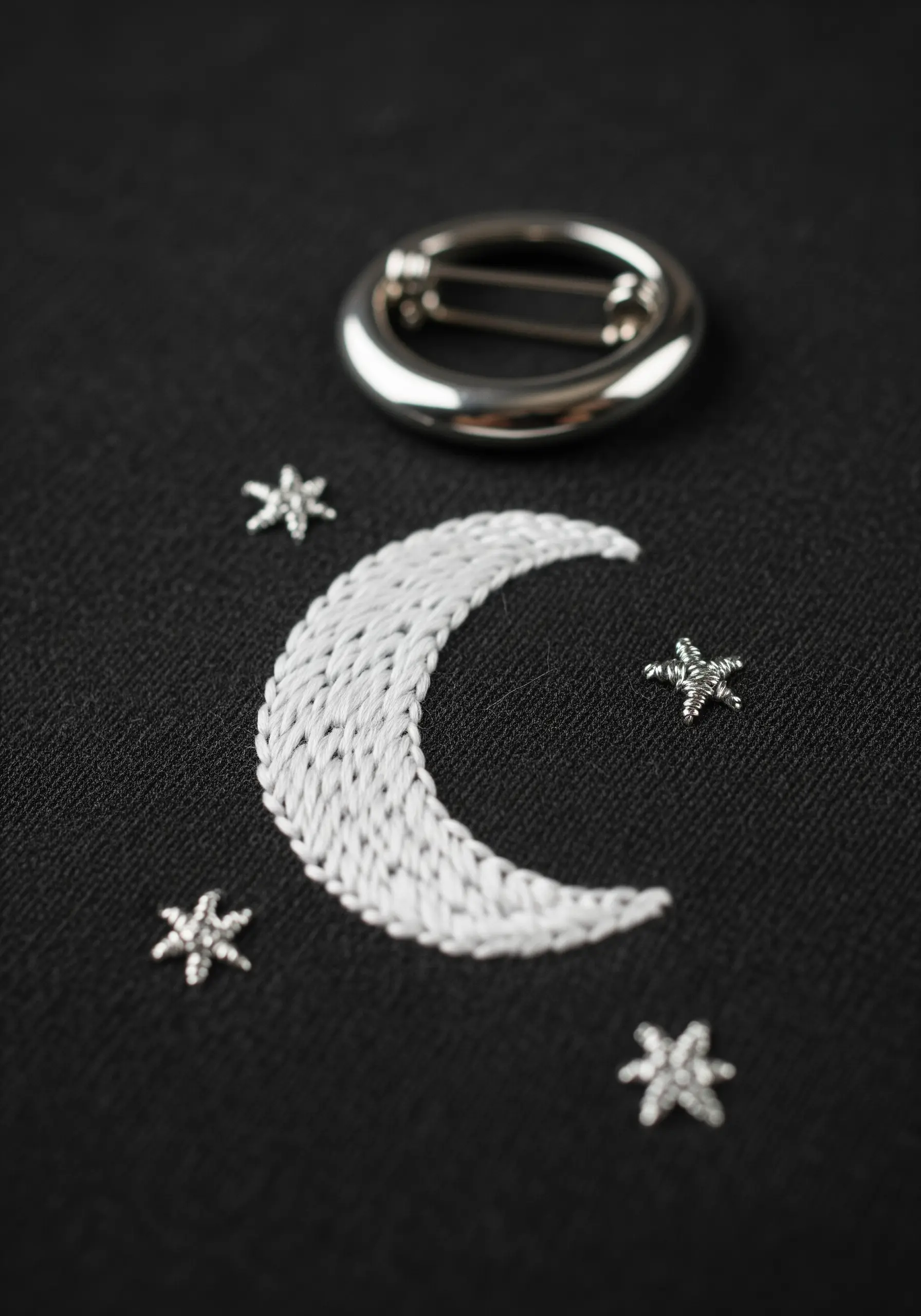 A white satin-stitched crescent moon with small silver star beads on black fabric.