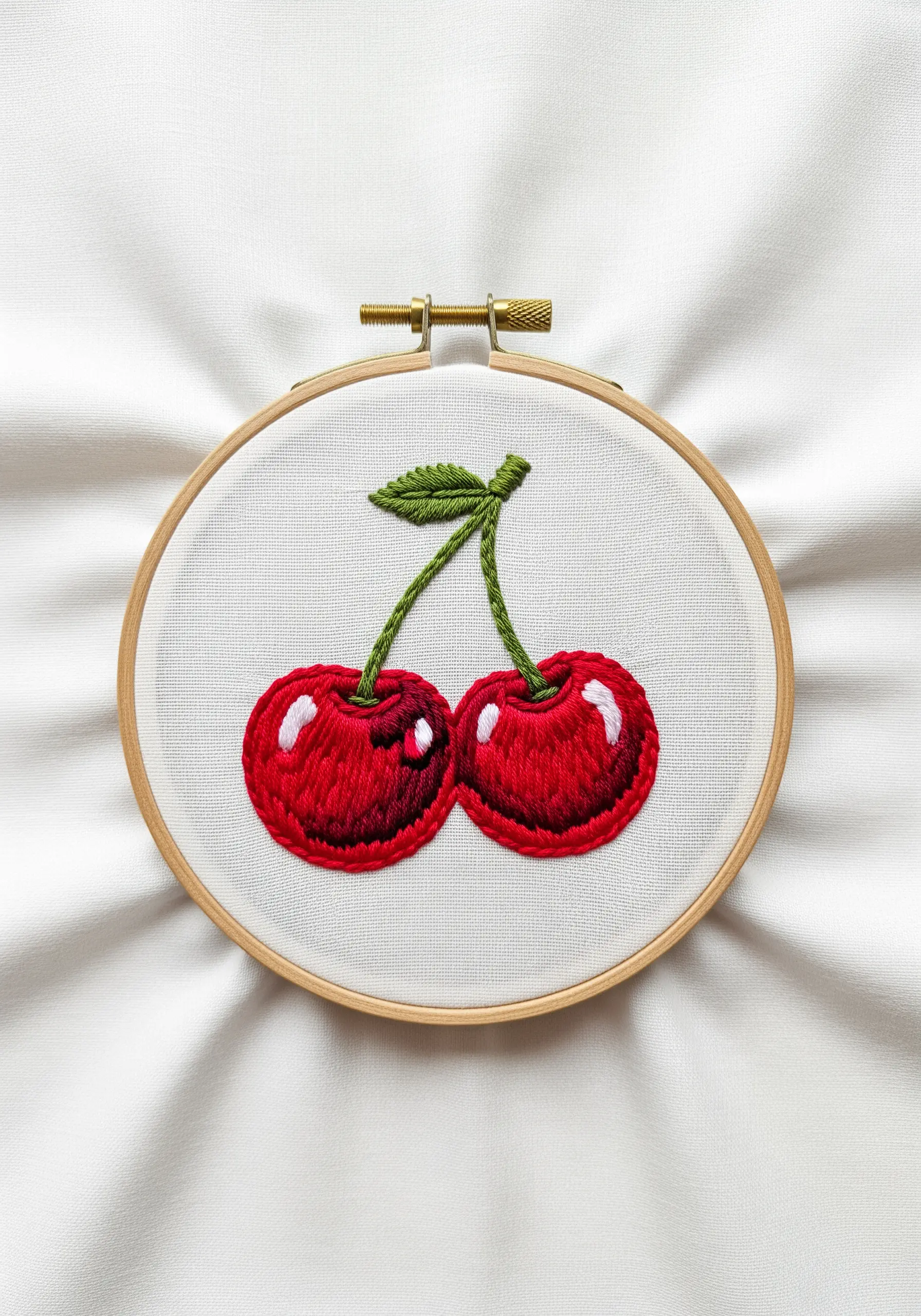 Two glossy, satin-stitched red cherries with bright white highlights in a hoop.