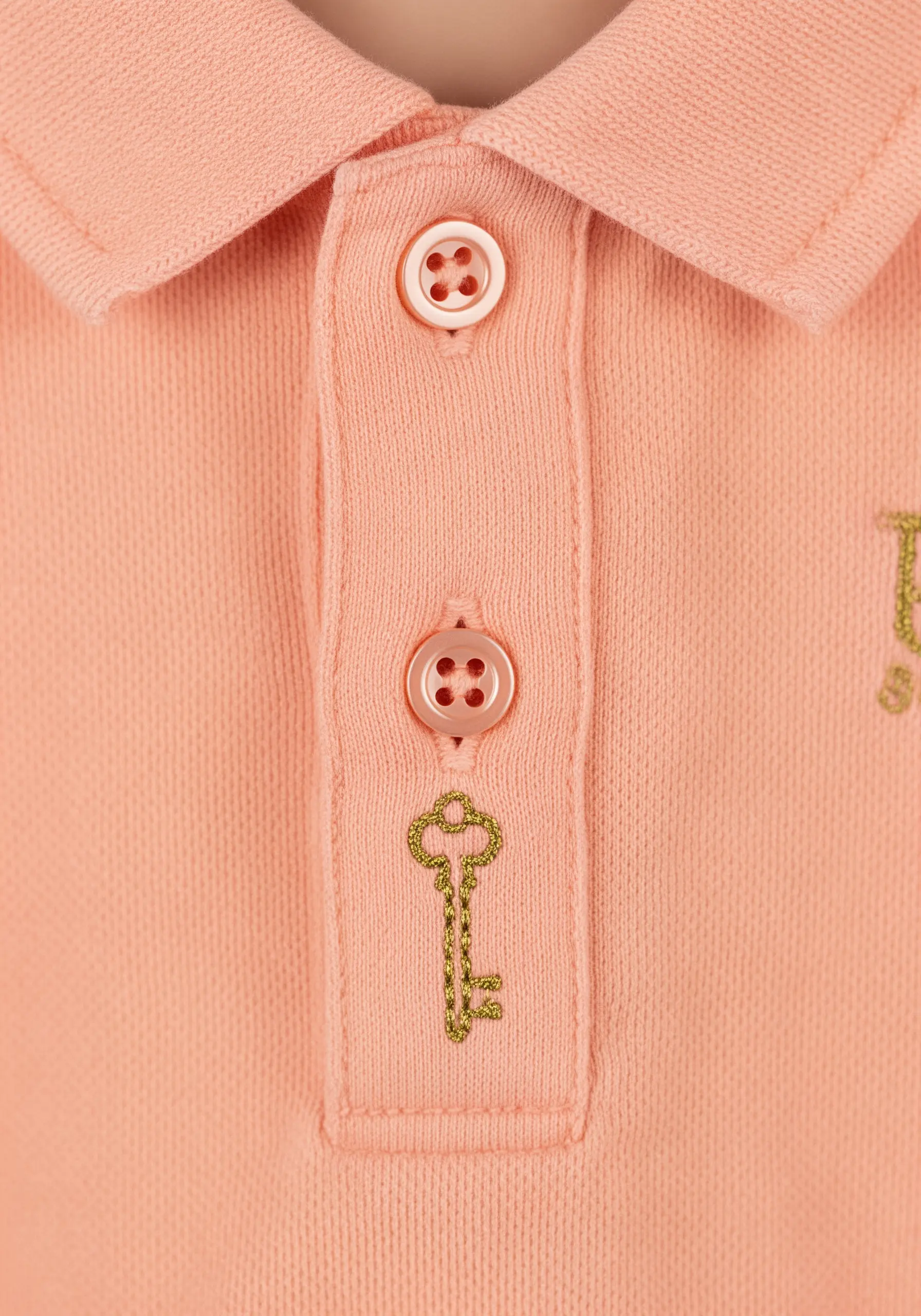 A small vintage key embroidered with gold metallic thread on a pink polo shirt.