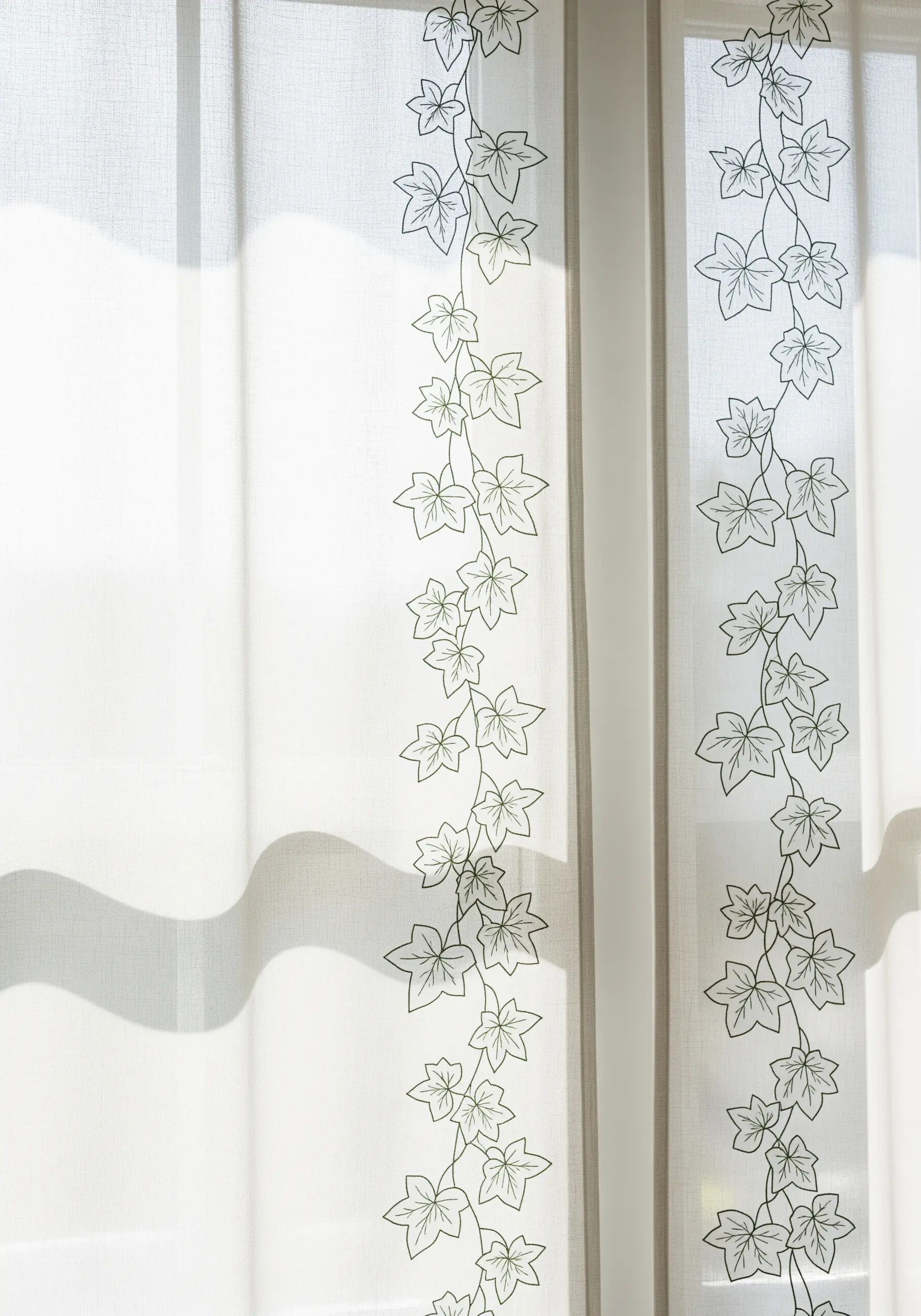 Sheer white curtains with a delicate, continuous vine and leaf pattern embroidered vertically.