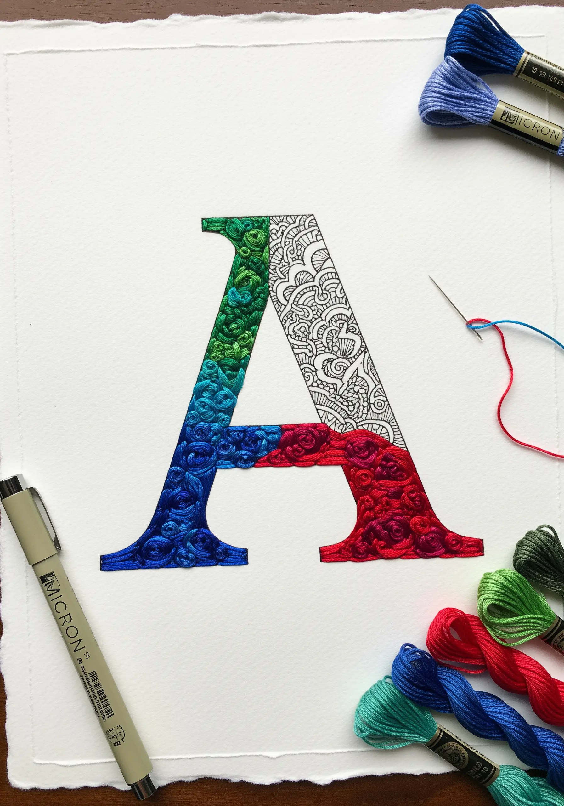 A large letter 'A' on paper, half filled with intricate ink patterns and half with colorful, dense embroidery.