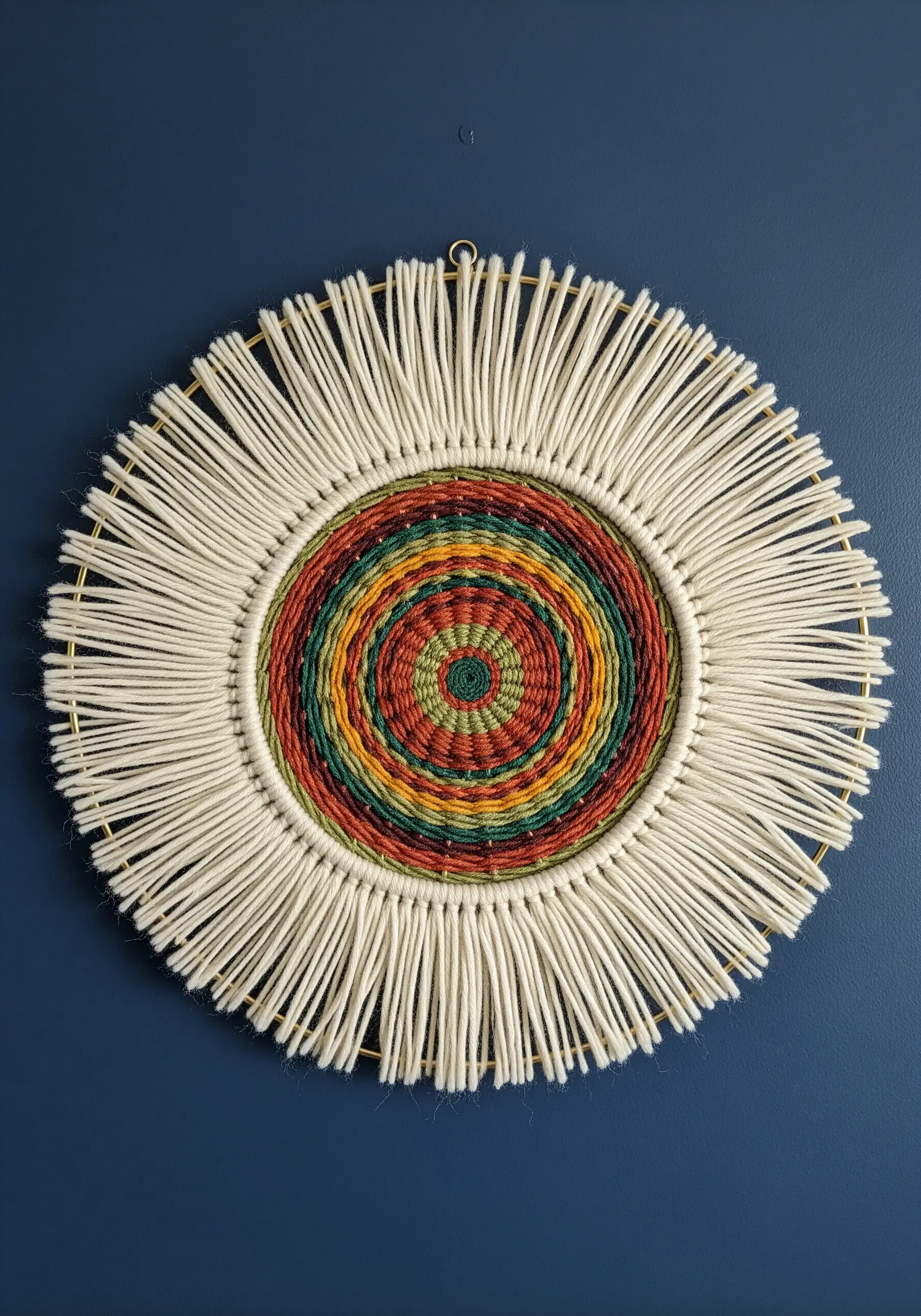 Circular weaving on a gold hoop with a colorful center and a natural yarn fringe.