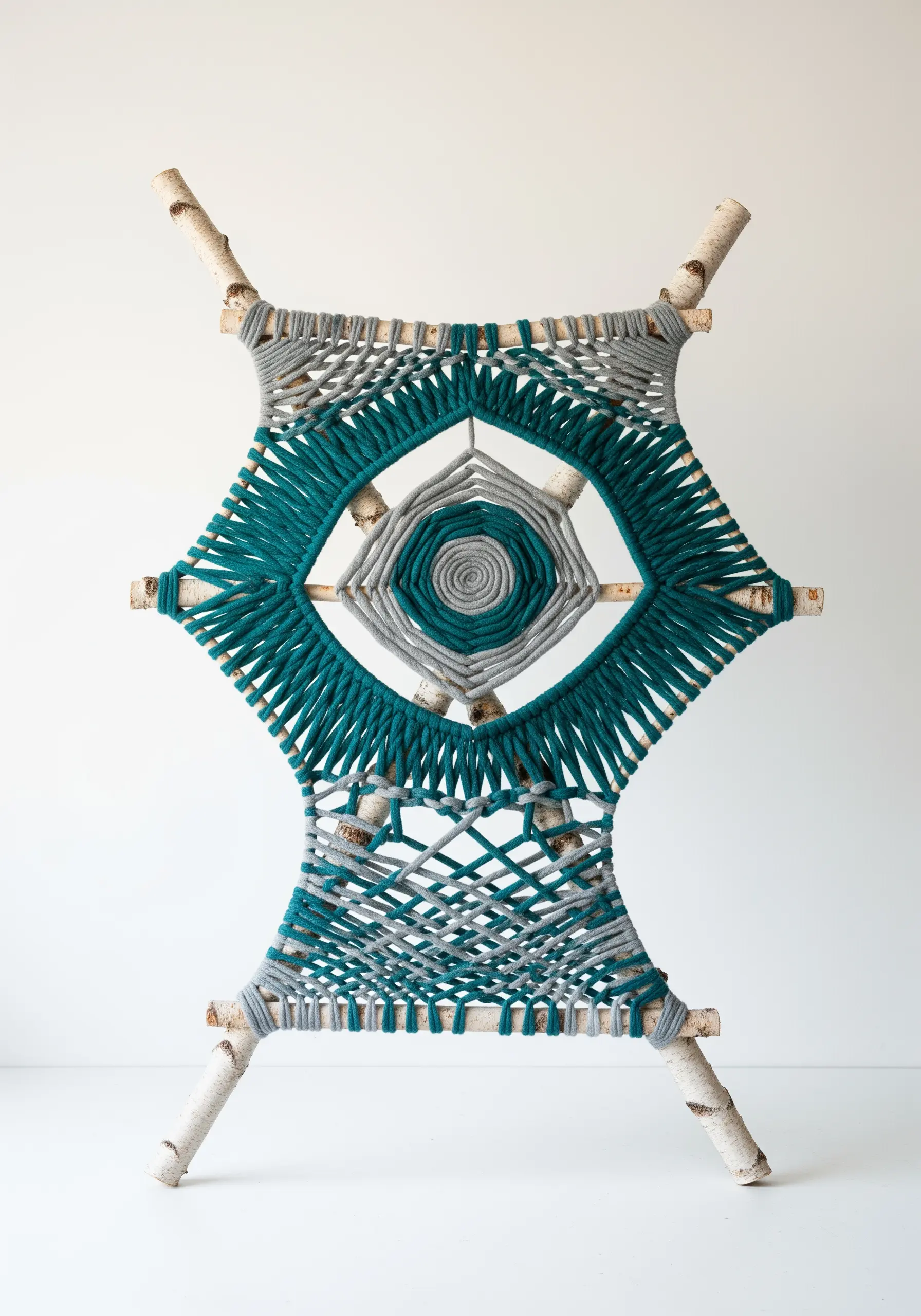 A sculptural yarn weaving in teal and gray wrapped around a frame of birch branches.