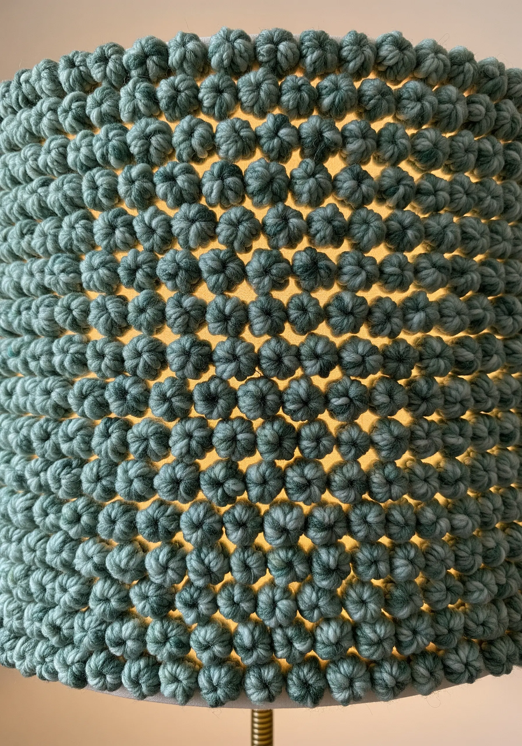 A close-up of a lampshade covered in densely packed green yarn knots, lit from within.