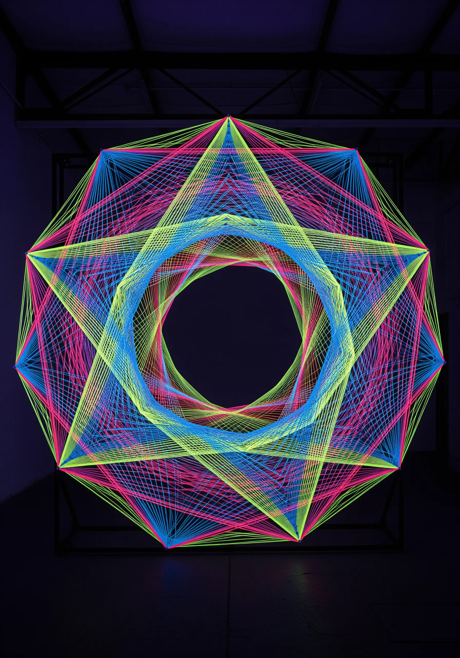 A geometric string art design using neon pink, blue, and yellow threads in a starburst pattern.