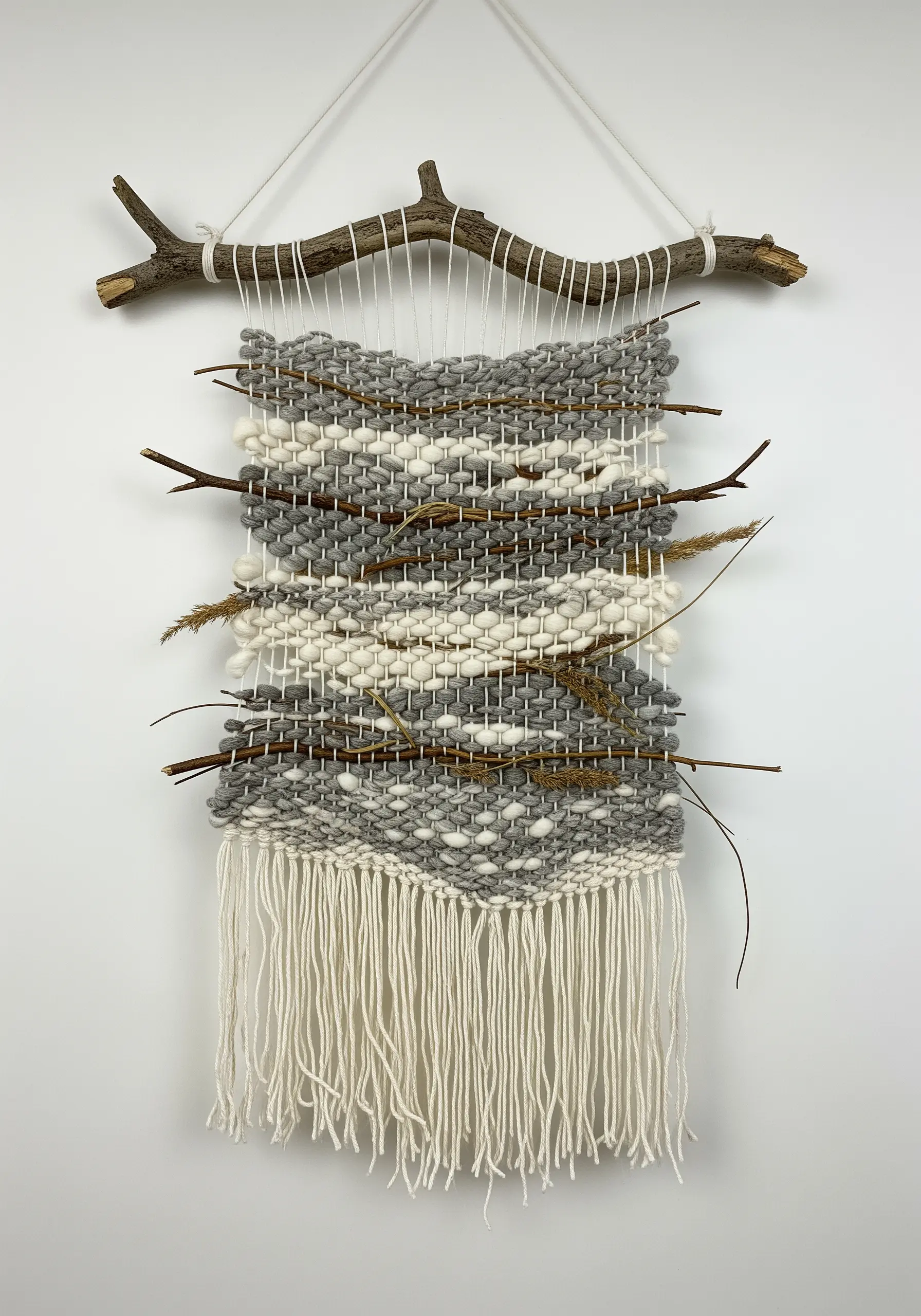 A small woven wall hanging in neutral tones, with small branches and dried grasses woven into it.