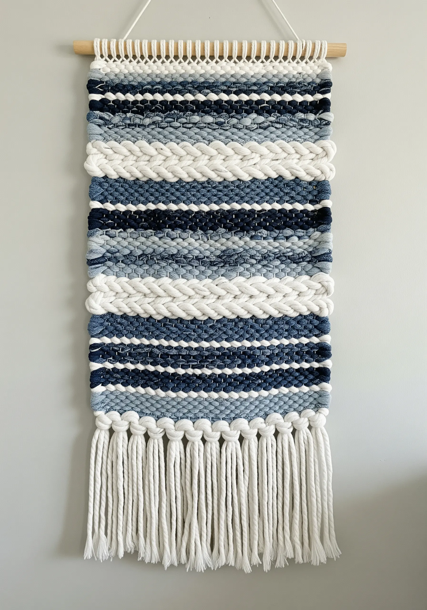 Woven wall hanging with horizontal stripes of blue yarn, white rope, and recycled denim.