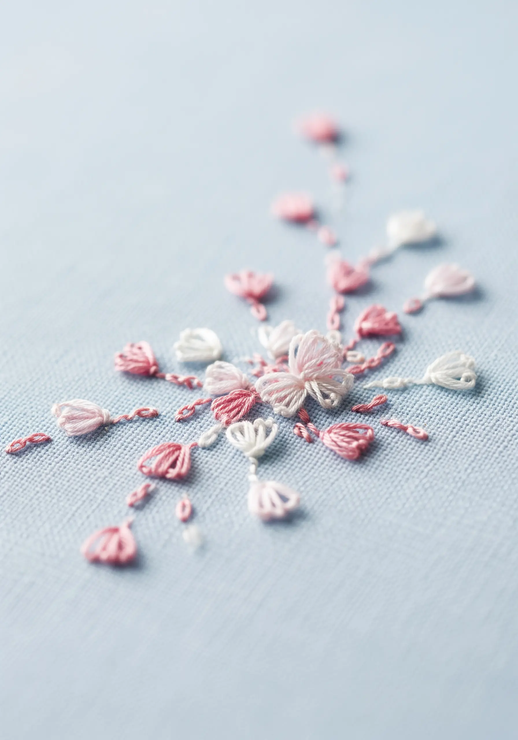 Delicate pink and white looped stitches forming tiny, abstract flowers on light blue fabric.
