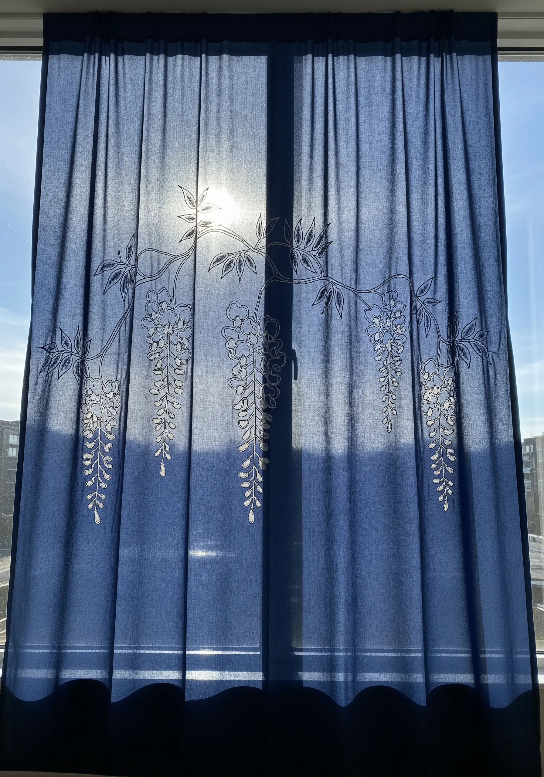 White outline embroidery of wisteria on a dark blue sheer curtain against a bright window.